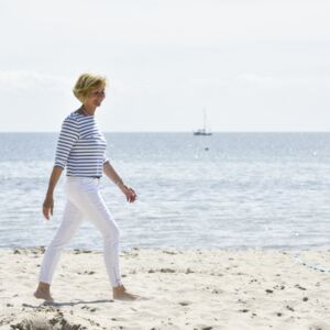 Eine Frau geht am Strand barfuss spazieren und sieht entspannt und glücklich aus. Im Hintergrund, auf der Ostsee, sind zwei Segelboote zu sehen.